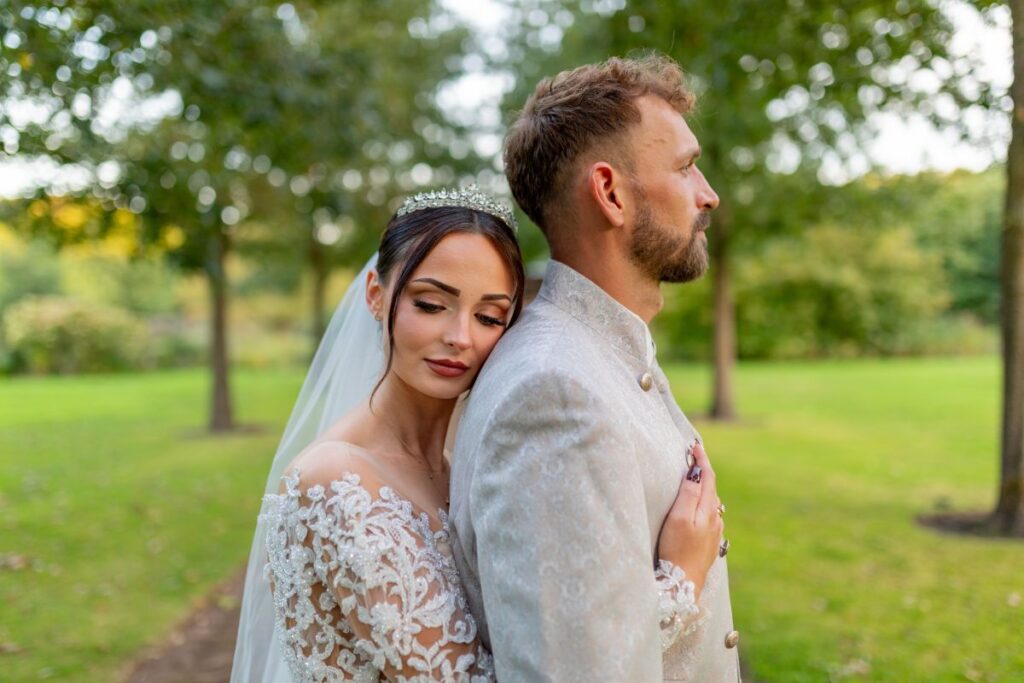 Braut und Bräutigam stehen in einem Park, Braut trägt ein elegantes Hochzeitskleid mit Spitze.
