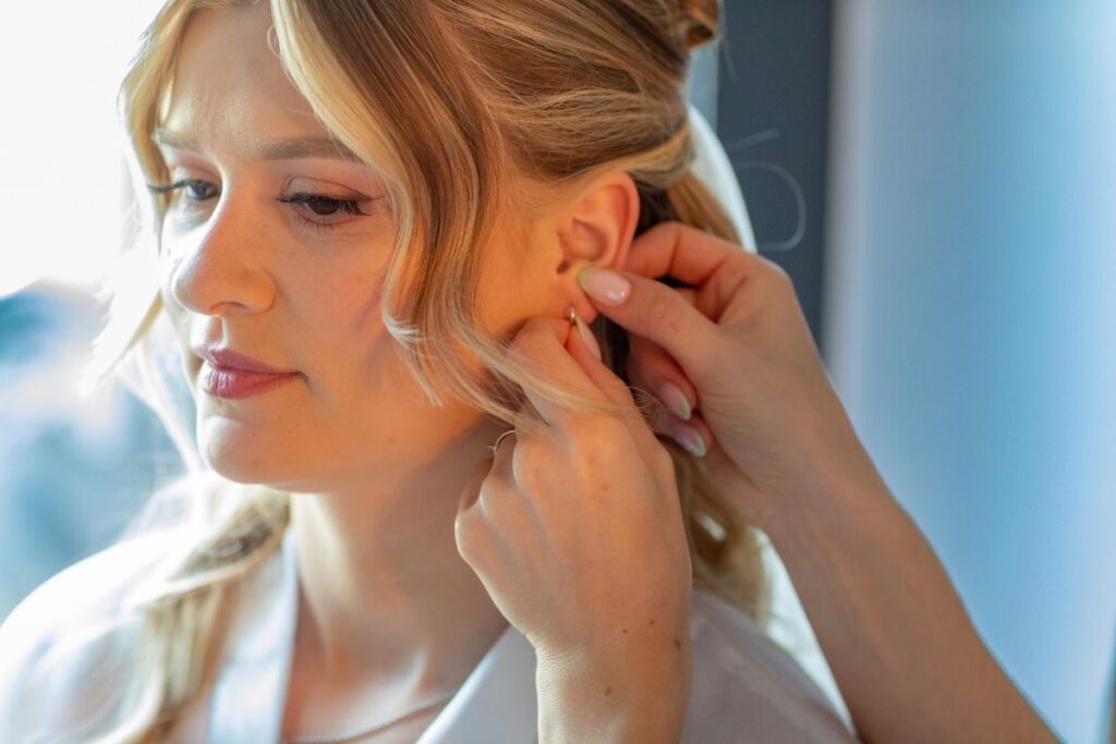 Getting Ready der Braut beim Anlegen der Ohrringe im natürlichen Fensterlicht