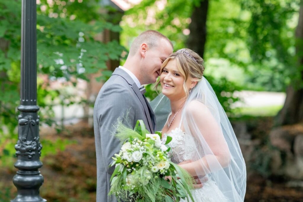 Braut und Bräutigam stehen eng beieinander im Grünen, die Braut lächelt in die Kamera