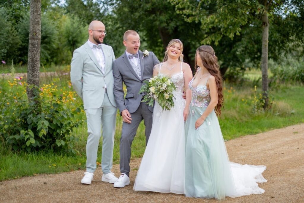 Brautpaar mit zwei Hochzeitsgästen steht lachend auf einem Weg in einer grünen Parklandschaft