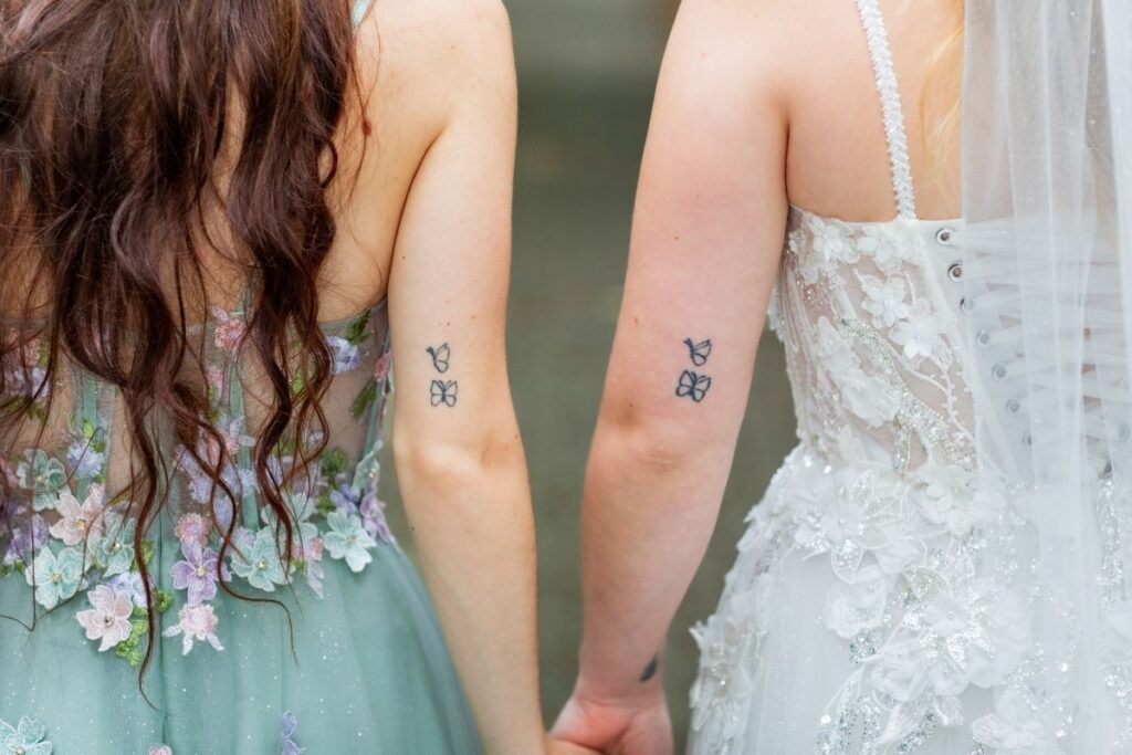 Zwei Frauen halten sich an den Händen, sichtbar sind passende Schmetterlings-Tattoos an den Armen
