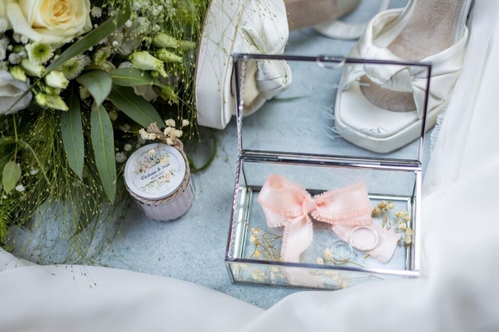 Detailaufnahme von Eheringen in einer Glasbox mit Schleife neben Brautschuhen und Blumen