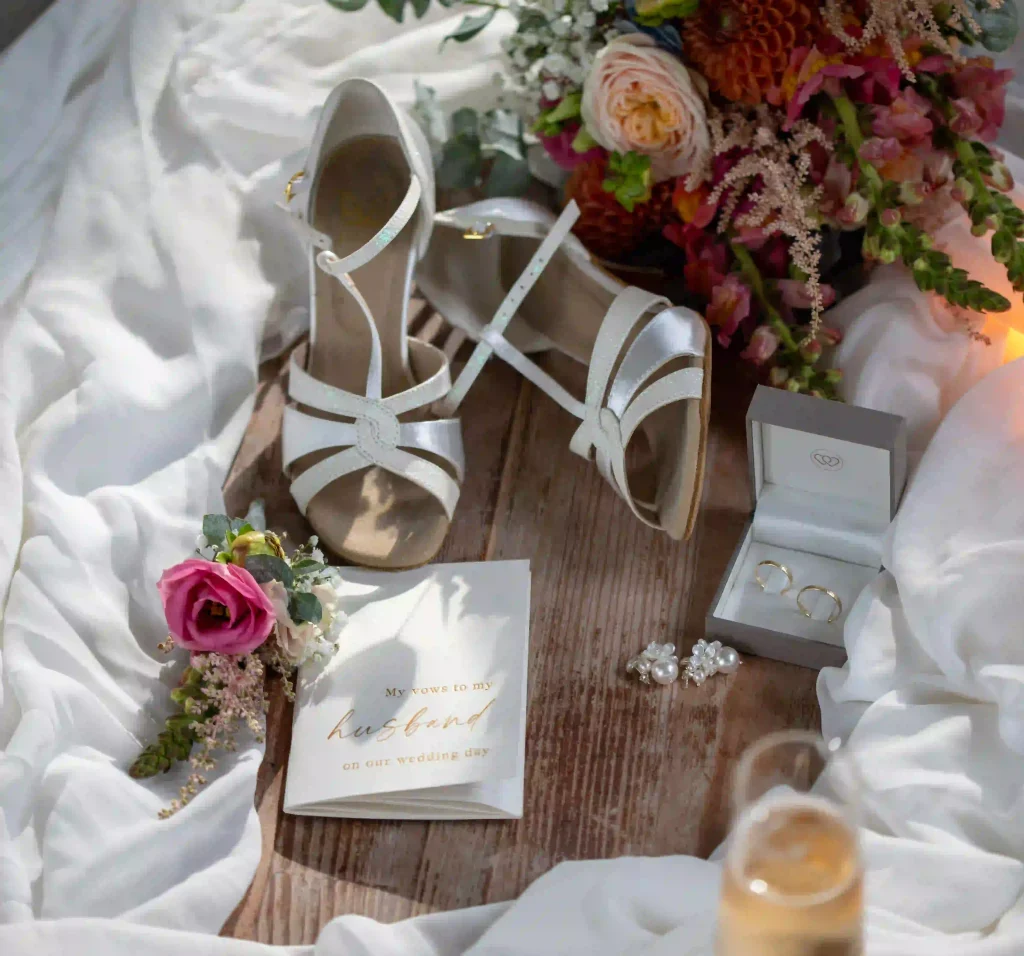 Flatlay mit weißen Brautschuhen, goldenen Eheringen, Brautschmuck und Blumenstrauß