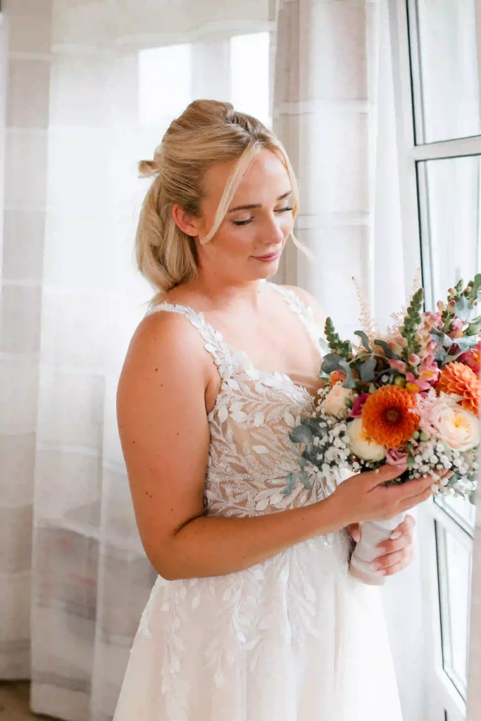 Braut im weißen Spitzenkleid betrachtet ihren farbenfrohen Brautstrauß beim Hochzeitsfotoshooting