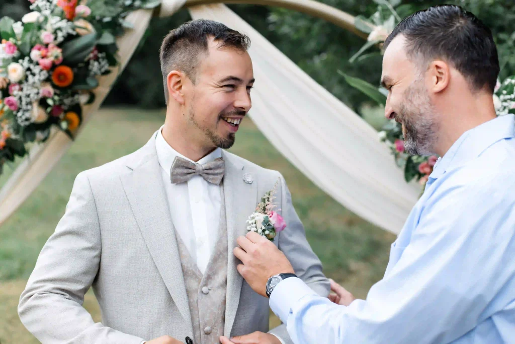 Hochzeit – Bräutigam wird vor der Trauung der Blumenanstecker am hellgrauen Anzug gerichtet