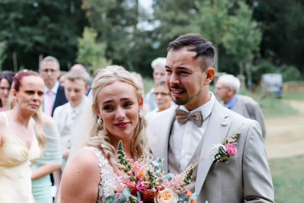 Brautpaar bei der Hochzeit im Freien – Braut mit buntem Blumenstrauß und Bräutigam im hellgrauen Anzug. Das Brautpaar weint vor Freude.