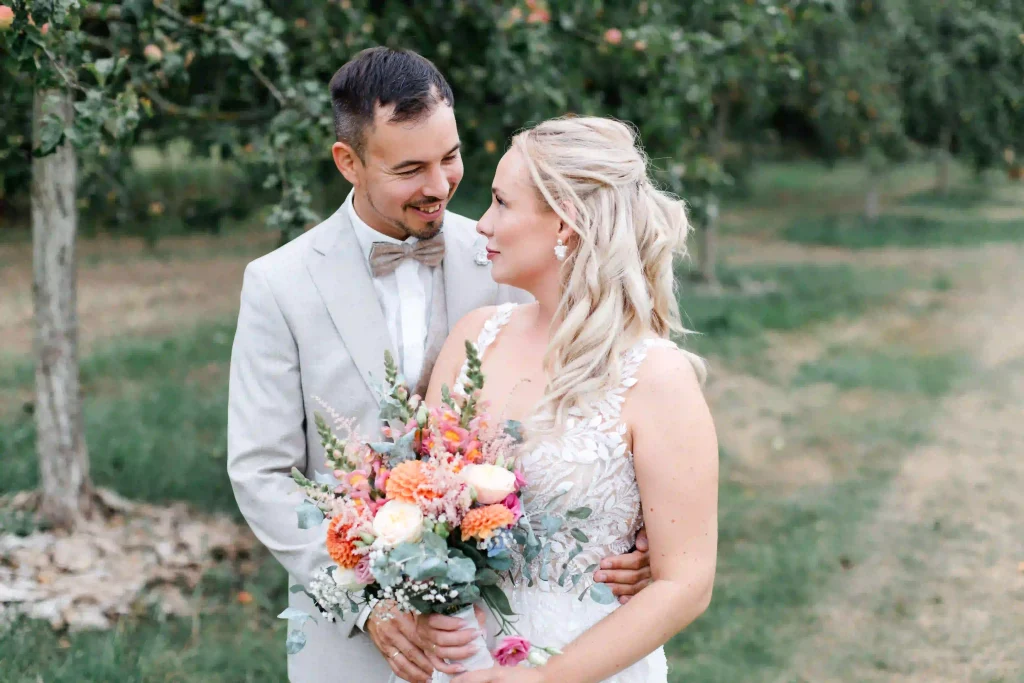 Braut und Bräutigam lächeln sich bei ihrem Hochzeitsportrait liebevoll an und halten einen bunten Blumenstrauß.